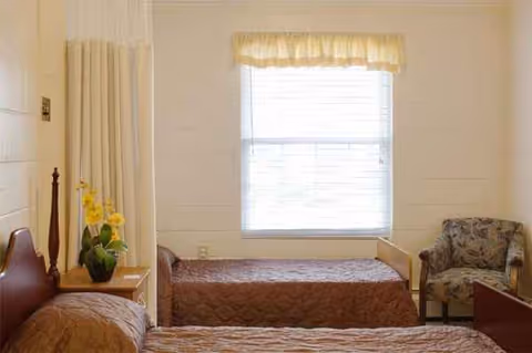 Two twin beds in a small, cozy bedroom with a window and chair.