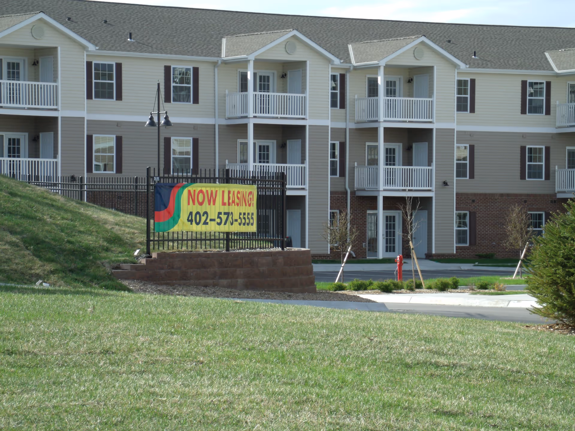 Exterior view of a multi-story residential building with balconies and white railings. In front of the building, there is a black metal fence with a yellow sign that reads 'NOW LEASING! 402-573-5555'. The foreground shows a grassy area and a paved driveway.