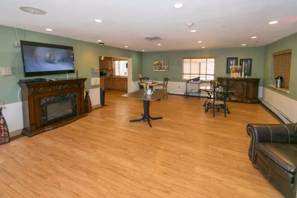A spacious common area with wooden flooring, green walls, and recessed ceiling lights. The room features a wall-mounted flat-screen TV above a decorative wooden fireplace, several small tables with chairs, a leather armchair, and a wooden cabinet with beverage dispensers. There is a window with blinds and an open doorway leading to a kitchen area.