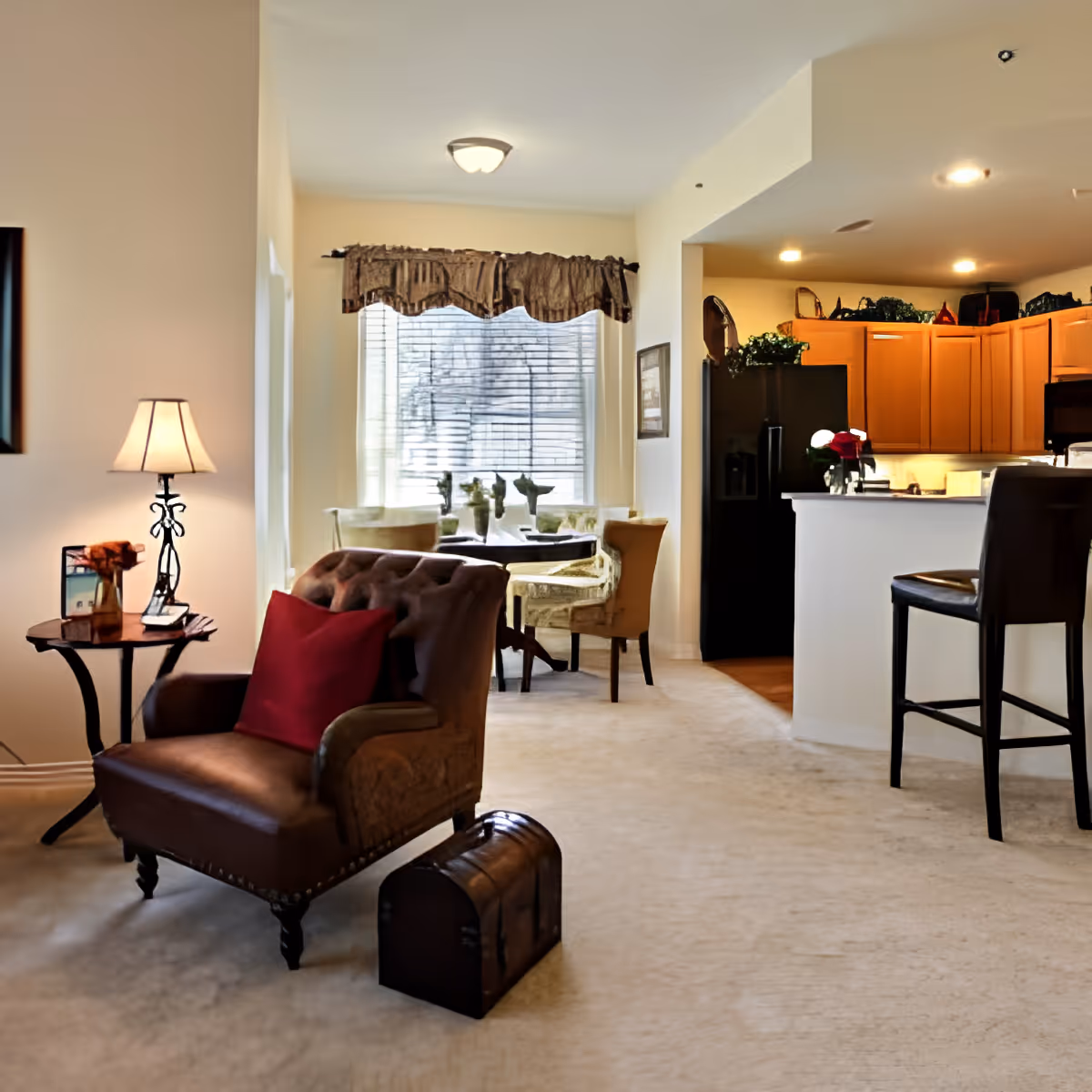 A cozy living area in a senior living facility featuring a brown tufted armchair with a red pillow, a small side table with a lamp and decorative items, a dining area with a round table and chairs near a window with blinds and a valance, and a kitchen with wooden cabinets, a black refrigerator, and a breakfast bar with a high chair.