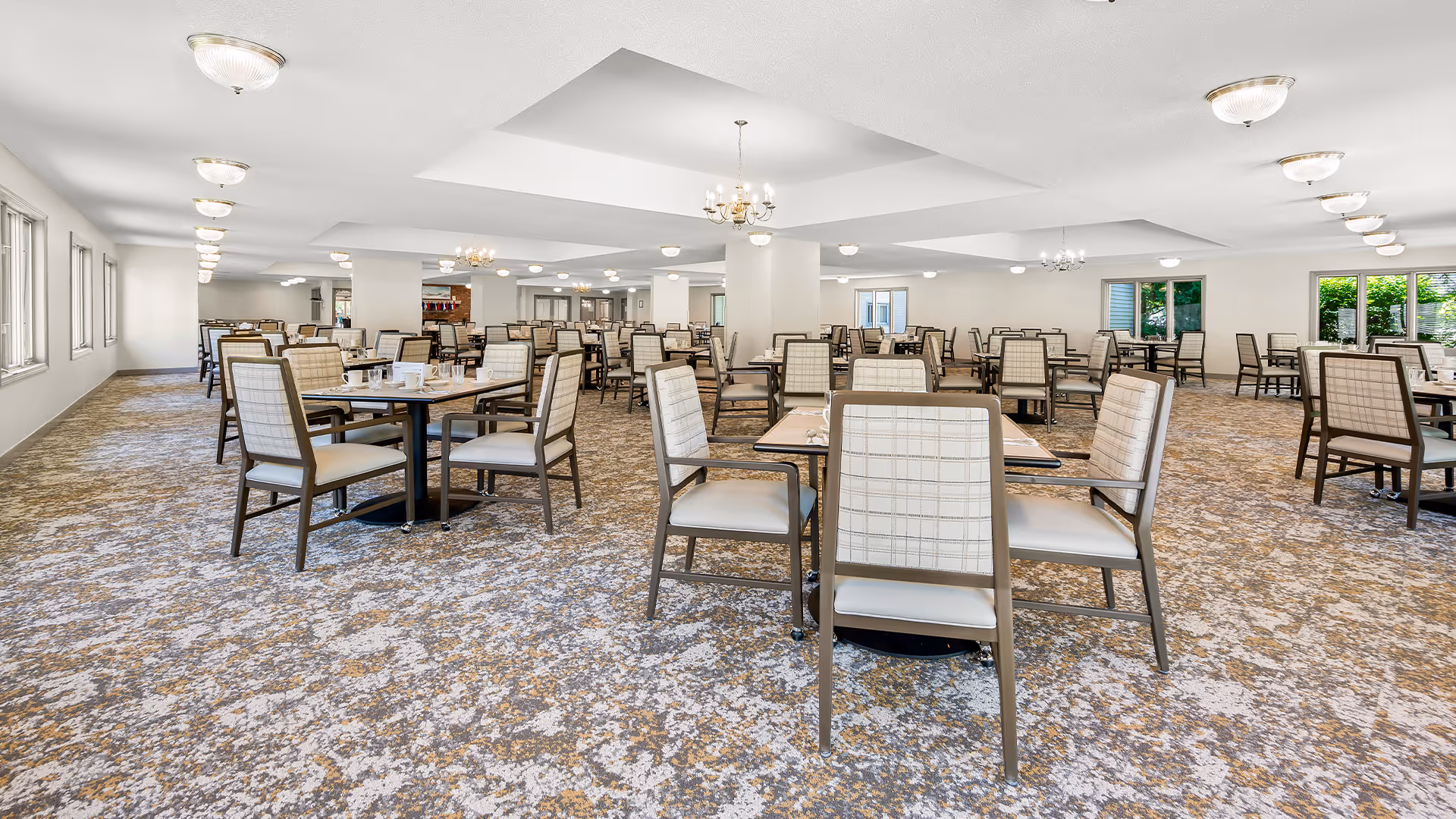 Large, bright dining room with many tables and chairs arranged on patterned carpet and chandeliers overhead.