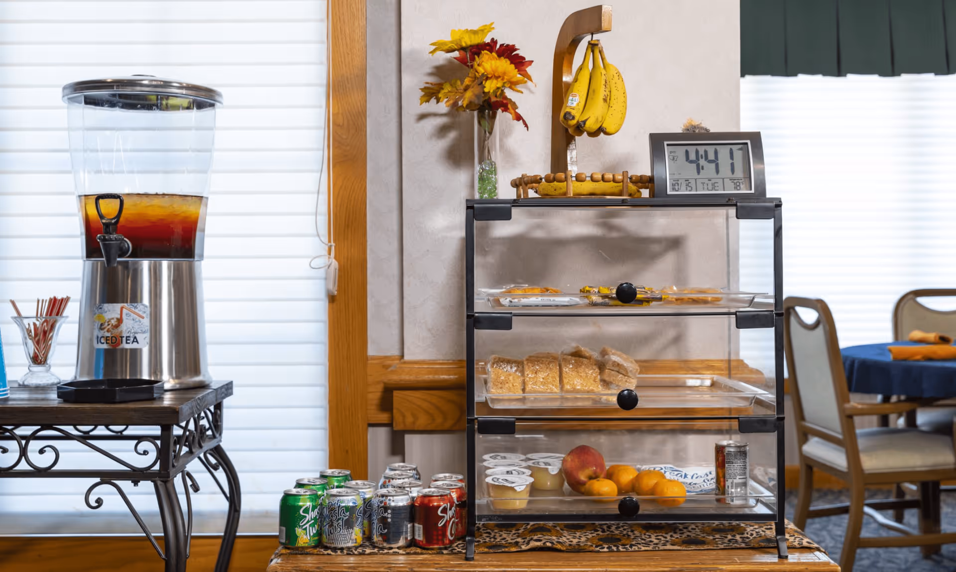A beverage station with a large iced tea dispenser on a metal table, several cans of soda on the floor, and a three-tiered clear display case containing bananas, bread, fruit cups, and oranges. A digital clock showing 4:41 is placed on top of the display case, and a vase with artificial flowers is also visible. In the background, there are dining chairs and tables with blue tablecloths and orange napkins.