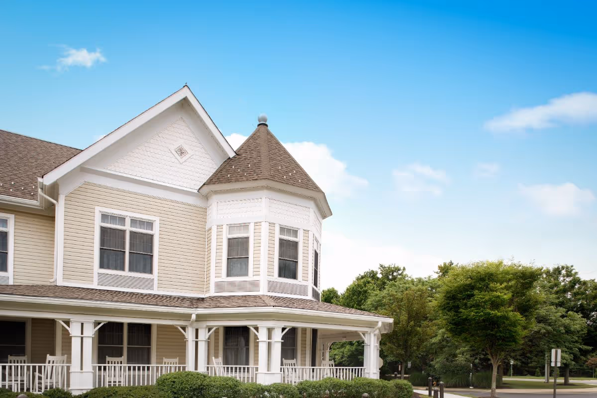 Front exterior of a light-colored two-story building with a wraparound porch and turret under a blue sky.