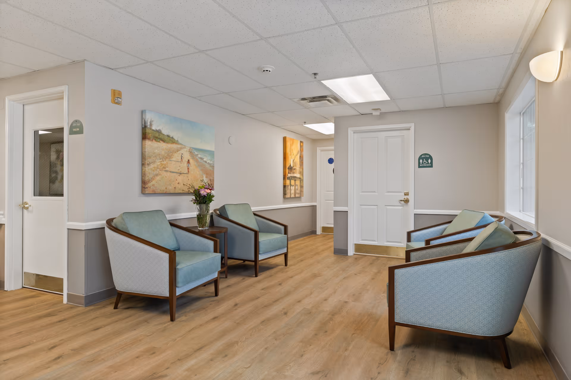 A bright seating area in a memory care facility with several upholstered armchairs, side tables, wall art, and white doors.