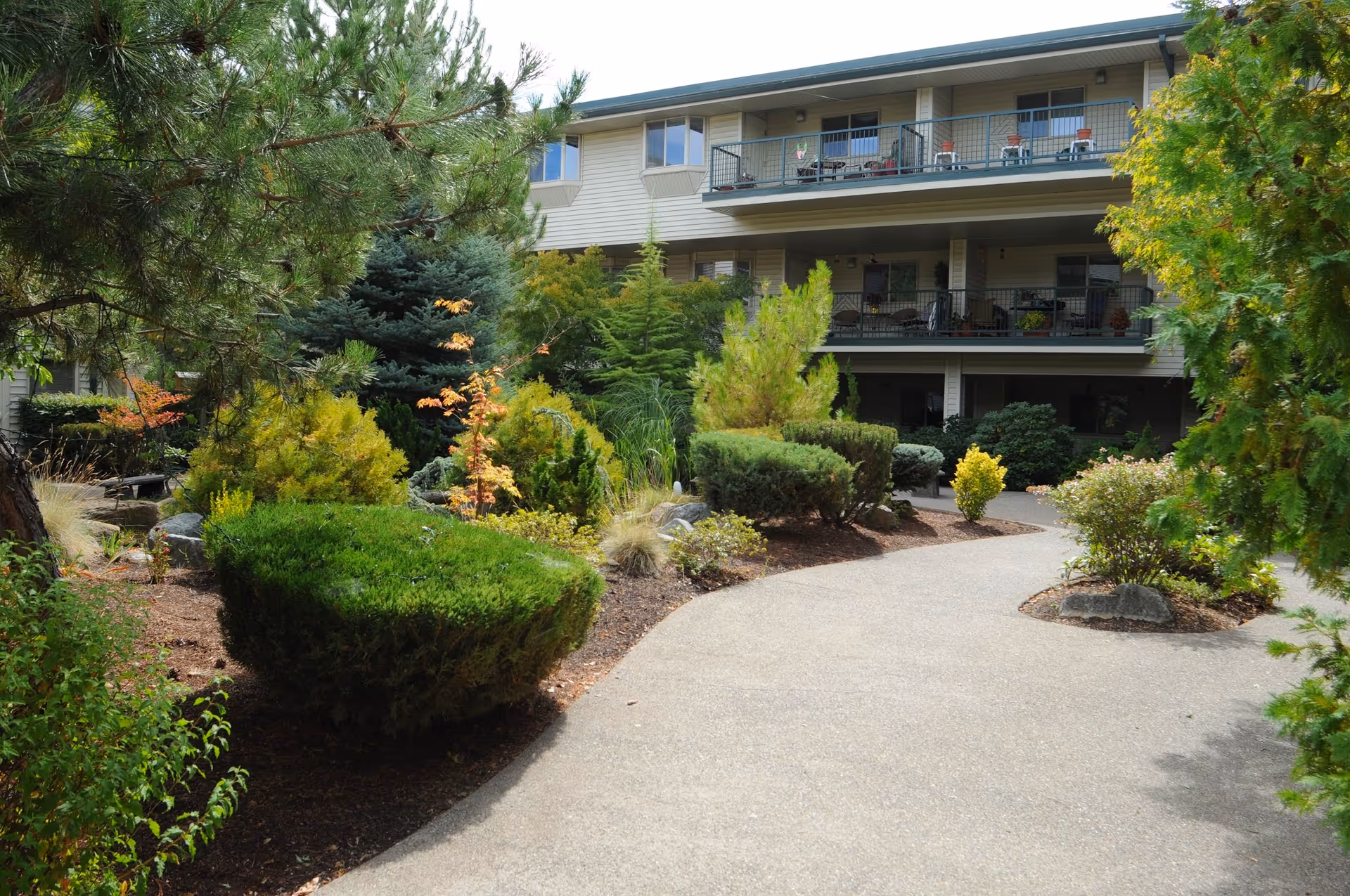 Curved walkway through a landscaped courtyard with shrubs and trees leading to a multi-story building with balconies.
