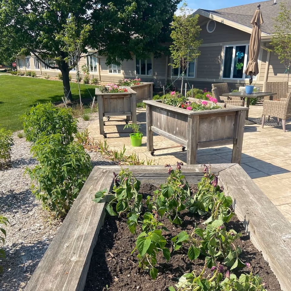 Outdoor patio area at The Marquardt featuring raised wooden garden beds with plants, a paved walkway, green lawn, trees, and a seating area with wicker chairs and a table with a closed umbrella near a beige building.