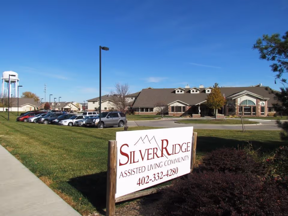 Exterior view of Silver Ridge Assisted Living Community building with a parking lot filled with cars, a water tower in the background, and a clear blue sky.