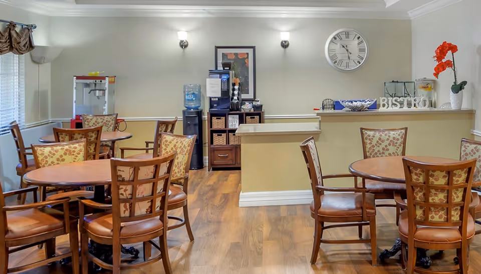 Interior view of a dining area with round wooden tables and cushioned chairs. The room has wood flooring, a water dispenser, a coffee machine on a cabinet, a popcorn machine, and a counter with decorative items including a clock on the wall and a vase with red flowers.