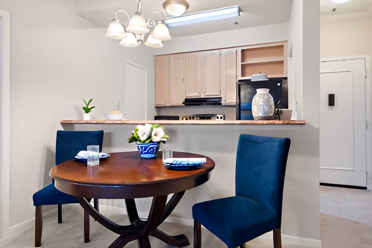 A small dining area with a round wooden table set for two with blue upholstered chairs. A blue vase with white flowers is on the table. Behind the dining area is a kitchen with light-colored cabinets, a black refrigerator, and a countertop with decorative items. A chandelier with multiple lights hangs above the table.