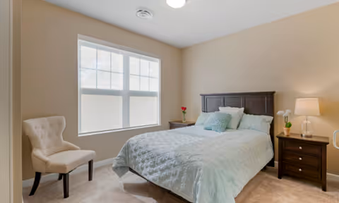 A bright bedroom with a made bed featuring light blue bedding, dark wood headboard and nightstands, a lamp, tufted chair, and a large window.