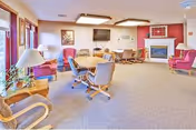 Lounge area with chairs, tables, fireplace, and a wall-mounted TV in a senior living facility.
