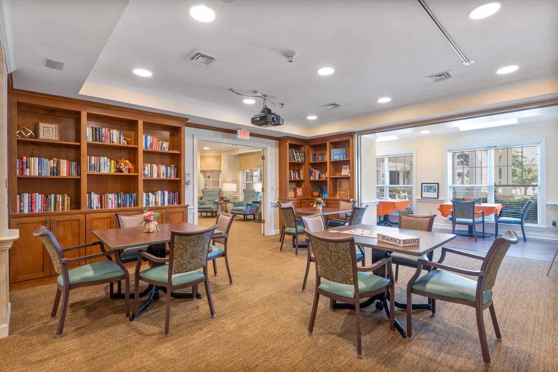 A bright and spacious common area in a senior living facility featuring multiple wooden tables with chairs arranged around them. The room has built-in wooden bookshelves filled with books and decorative items. There is a game of Sequence set up on one of the tables. Large windows allow natural light to fill the space, and an adjoining room with additional seating is visible through an open doorway.