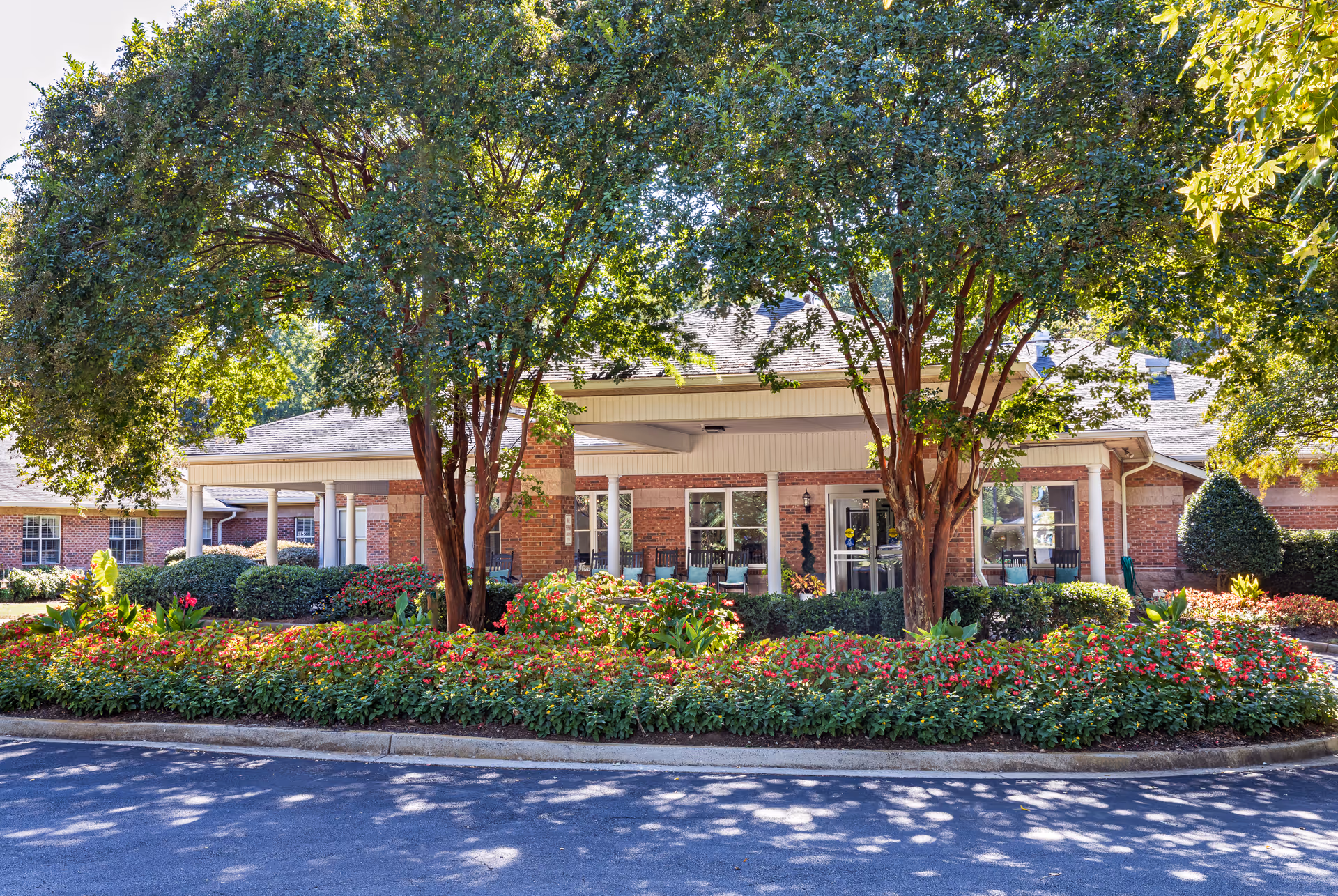 Front exterior view of Kempton of Rock Hill facility with a covered entrance, brick walls, large windows, and a landscaped garden with flowering plants and trees in front.