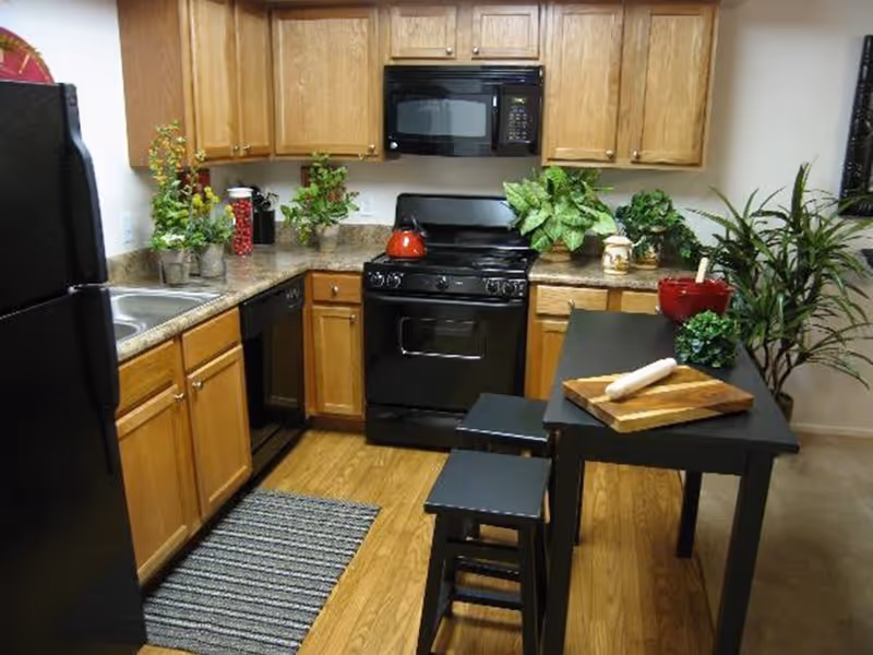 A kitchen with wooden cabinets, black appliances including a refrigerator, stove, microwave, and dishwasher. There is a black table with two stools, a cutting board with a rolling pin, and several green plants on the countertops and floor. The floor is wooden with a striped rug near the sink.