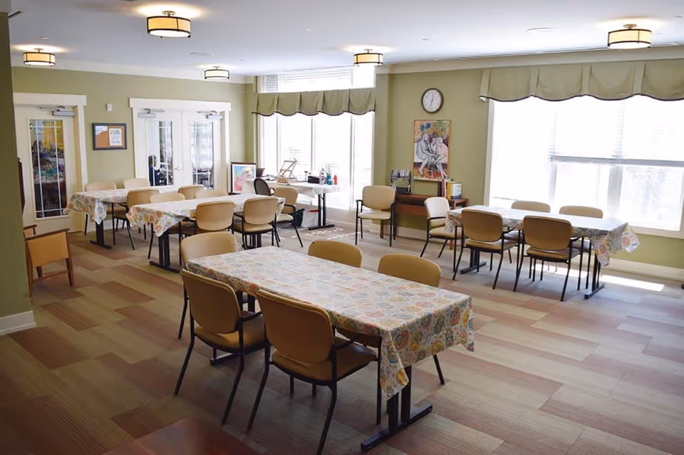 Communal dining room with multiple tables covered in patterned tablecloths, chairs, large windows, and soft green walls.