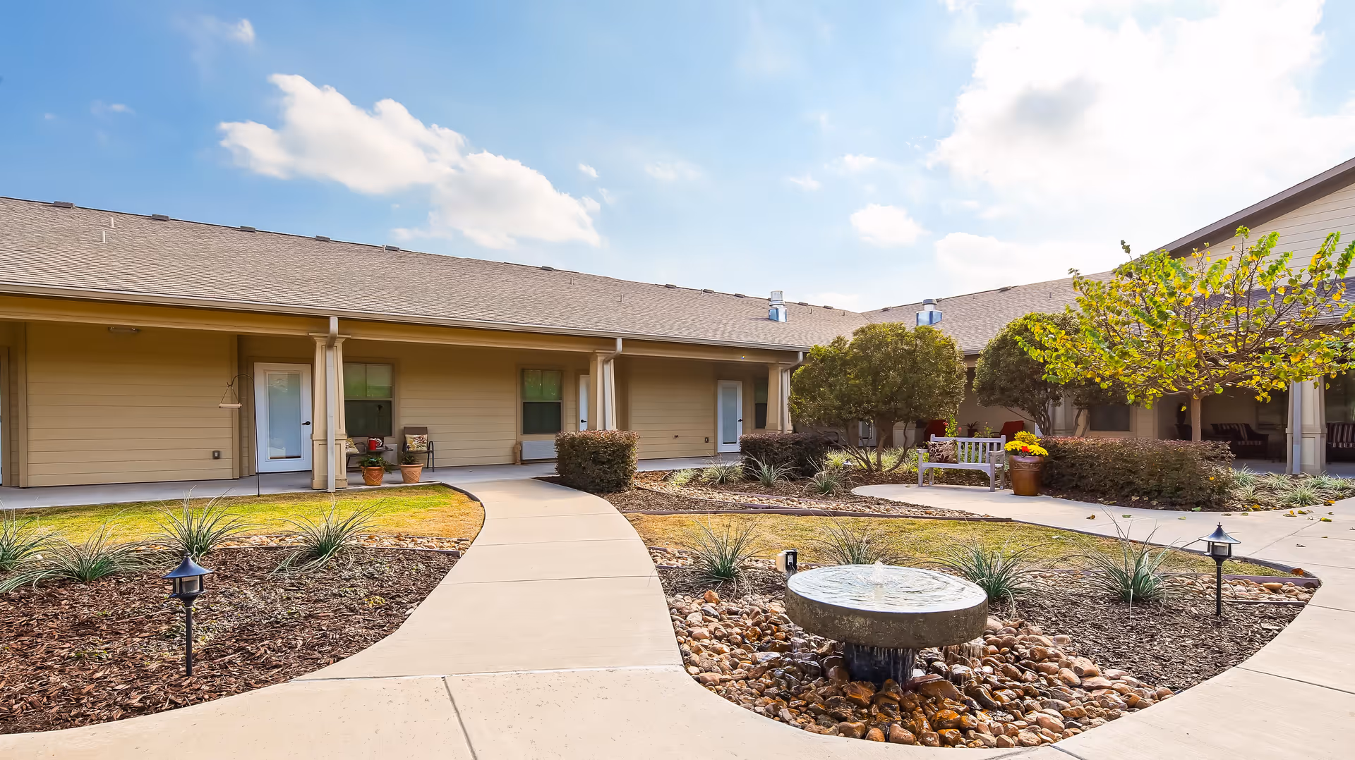 Outdoor courtyard area of Rock Ridge Assisted Living and Memory Care featuring a circular water fountain surrounded by rocks and plants, a curved concrete walkway, benches, potted plants, and a building with beige siding and a shingled roof under a partly cloudy sky.