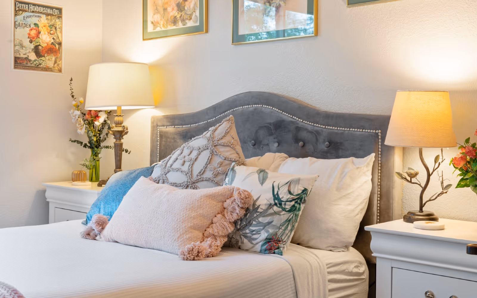 A cozy bedroom with a neatly made bed featuring a gray upholstered headboard adorned with nailhead trim. The bed has multiple decorative pillows in various textures and patterns. On either side of the bed are white nightstands, each with a table lamp emitting warm light. There are floral decorations on the nightstands and framed artwork hanging on the wall above the headboard.