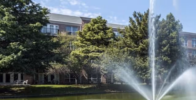 Brick multi-story building behind trees beside a pond with a central fountain spraying water.