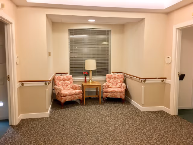 A small seating area in a hallway with two floral upholstered armchairs positioned on either side of a wooden side table with a lamp and a small vase with red flowers. The walls are beige with wooden handrails, and there is a window with closed blinds behind the chairs.