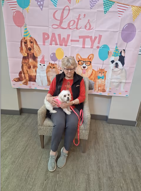 An elderly woman sitting on a chair holding a small white dog with a pink harness. Behind her is a colorful banner with illustrations of various dogs and cats wearing party hats and balloons, with the text 'Let's PAW-TY!'