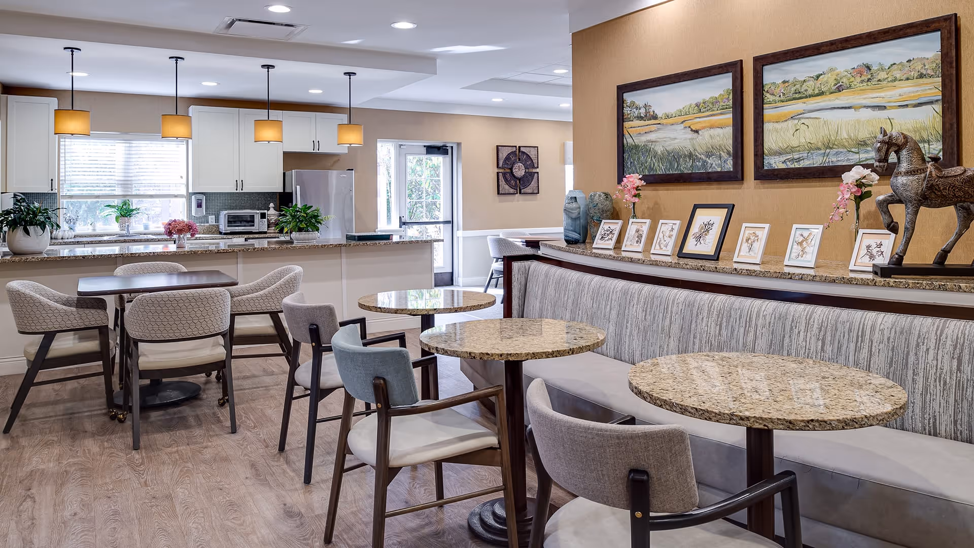 A bright and modern dining area in a senior living facility featuring round granite tables with upholstered chairs and a long cushioned bench along the wall. The background shows a kitchen with white cabinets, a granite countertop, and stainless steel appliances. The wall above the bench is decorated with framed artwork and small decorative items including a horse sculpture and flower arrangements.