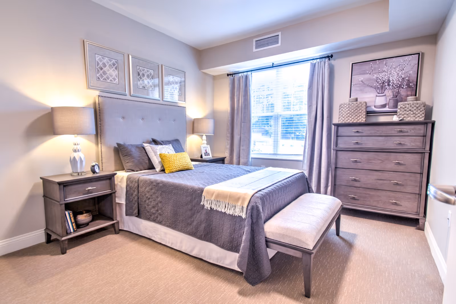 Well-lit furnished bedroom with a bed, bedside tables and a dresser in neutral tones.