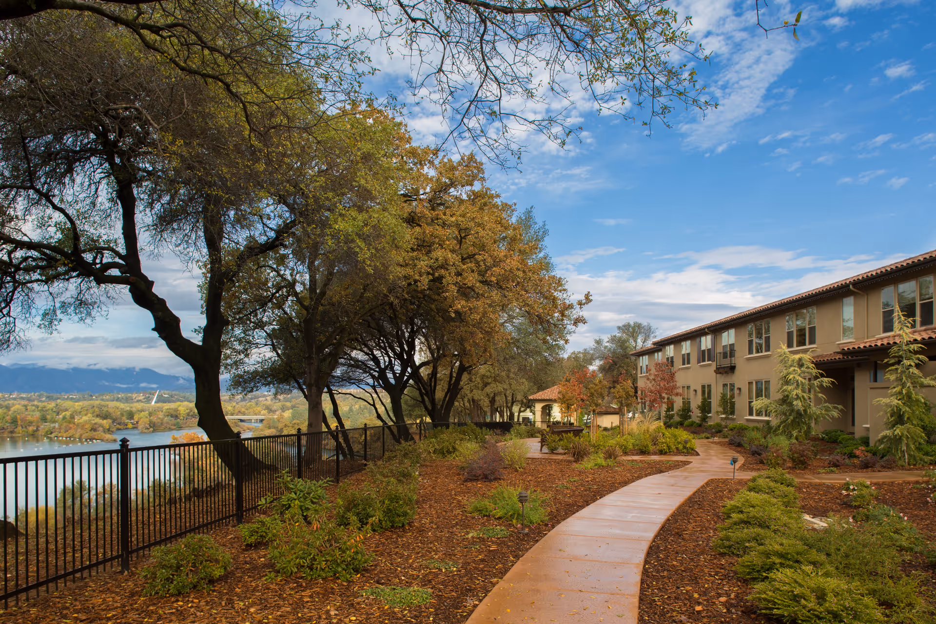 A landscaped outdoor walkway at Oakmont of Redding with a curved paved path, green shrubs, and trees. The path runs alongside a black metal fence overlooking a body of water and distant mountains under a partly cloudy sky. A two-story building with multiple windows is visible on the right side.