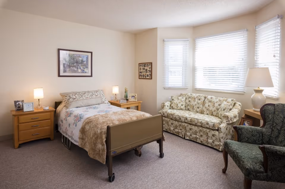 A cozy bedroom in a senior living facility featuring a single bed with floral bedding and a beige blanket. The room includes two wooden nightstands with lamps and framed photos, a floral-patterned sofa, a green upholstered armchair, and three windows with white blinds letting in natural light. A framed picture hangs on the wall above the bed.