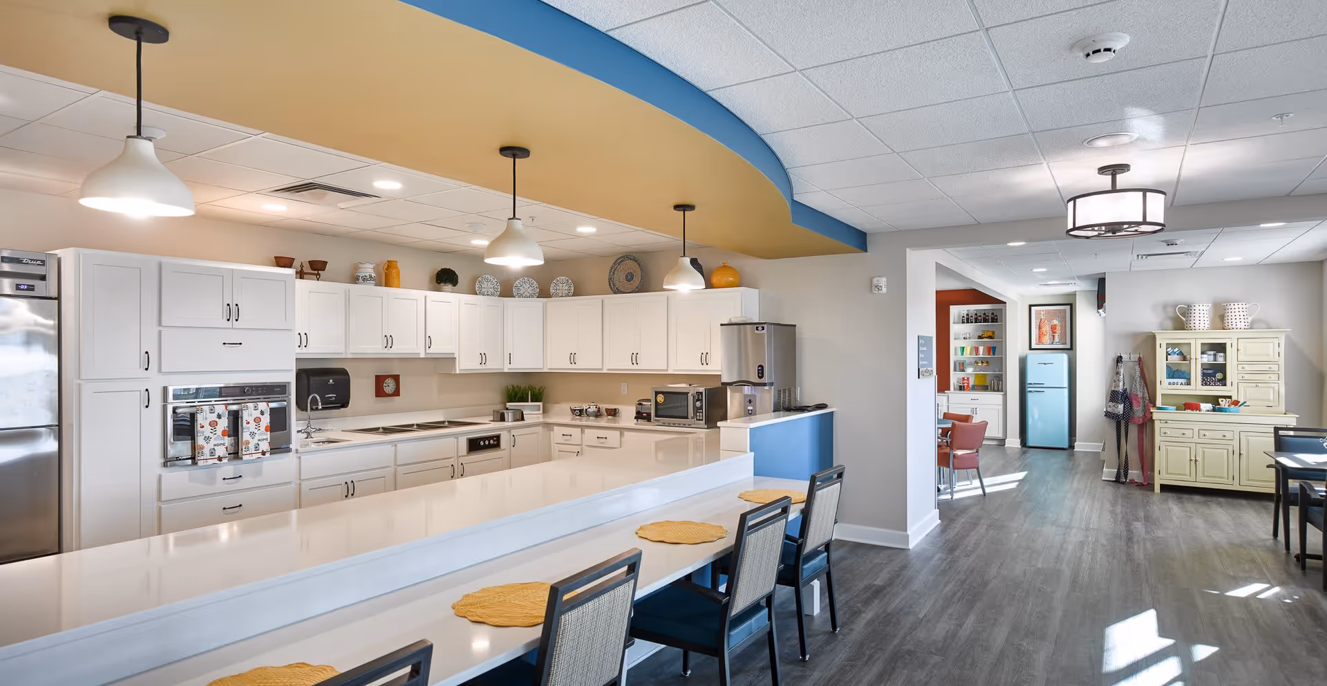 A bright, modern kitchen area in a senior living facility featuring white cabinetry, stainless steel appliances, and a long white countertop with four chairs and yellow placemats. The ceiling has a curved design with blue and yellow accents and three hanging pendant lights. In the background, there is a dining area with a light blue refrigerator, a yellow hutch, and several chairs and tables.