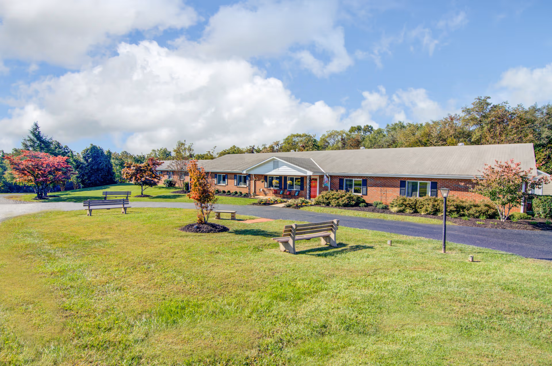 Exterior view of a single-story brick building with a gray roof, surrounded by green grass, trees with autumn foliage, and several benches on the lawn under a partly cloudy blue sky.