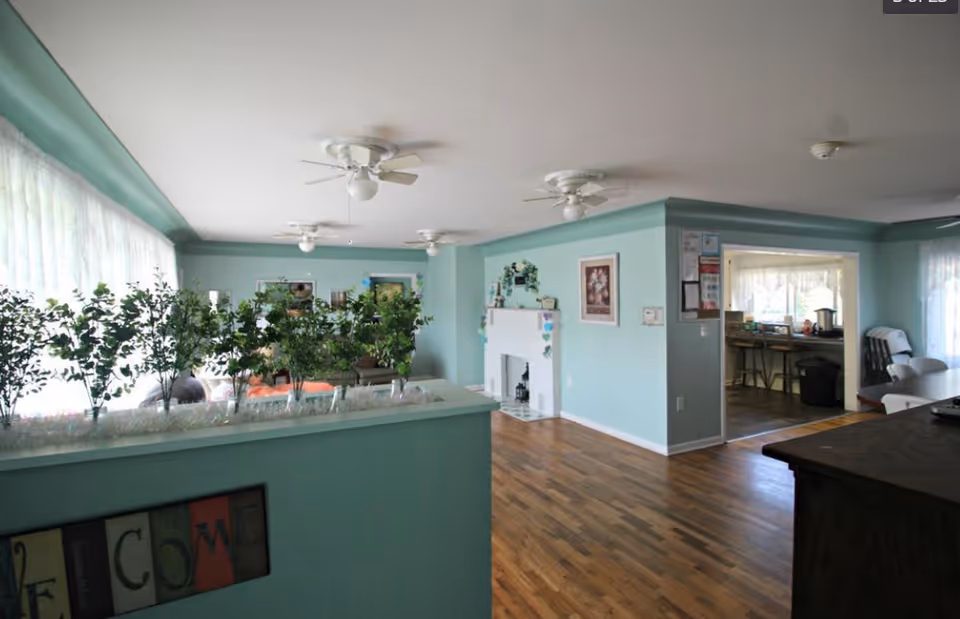 Interior view of a senior living facility common area with light blue walls, hardwood floors, ceiling fans, and a white fireplace. There are several green plants on a divider near large windows with sheer curtains. The room opens into a kitchen area with bar stools and a dining table with chairs.