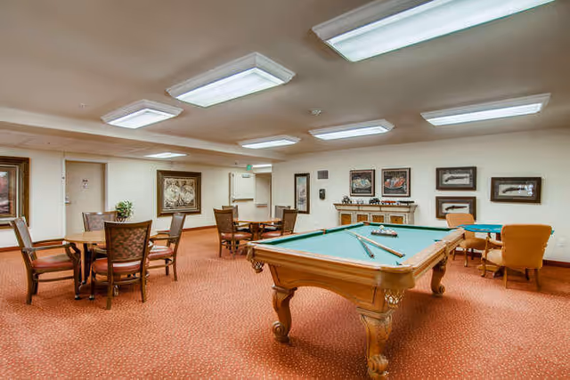 A spacious recreational room with a pool table in the center, surrounded by several wooden tables and chairs. The room has warm lighting from ceiling fixtures and framed artwork on the walls. The carpet is a reddish-brown color, and there are a few comfortable armchairs near the back wall.