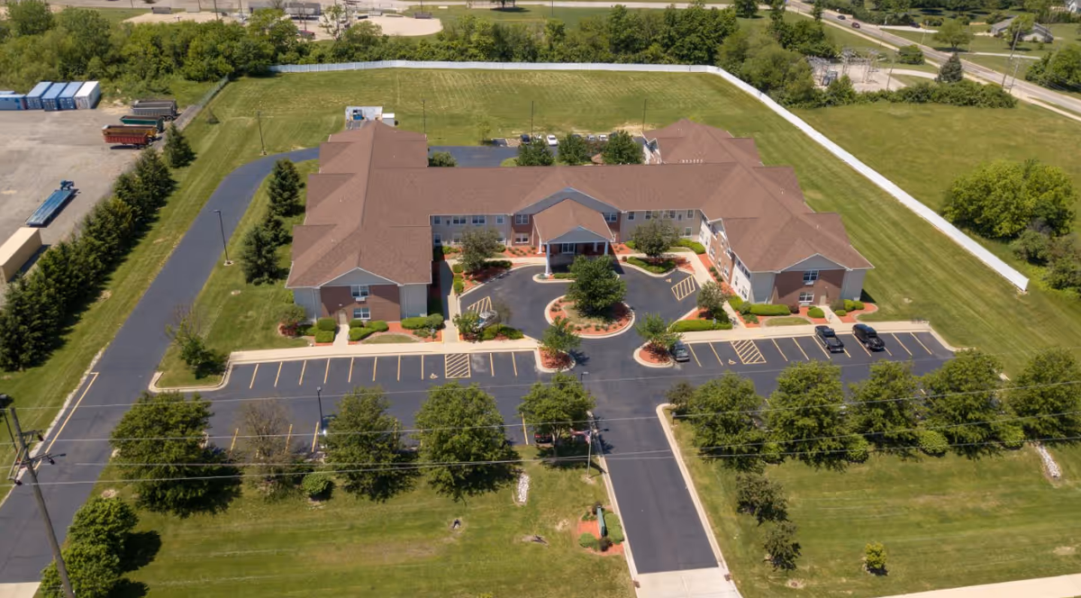 Aerial view of a single-story senior living facility building with a brown roof surrounded by green lawns and trees. The building has a circular driveway with parking spaces and landscaped areas with shrubs and small trees. The property is bordered by a white fence and adjacent to a road.