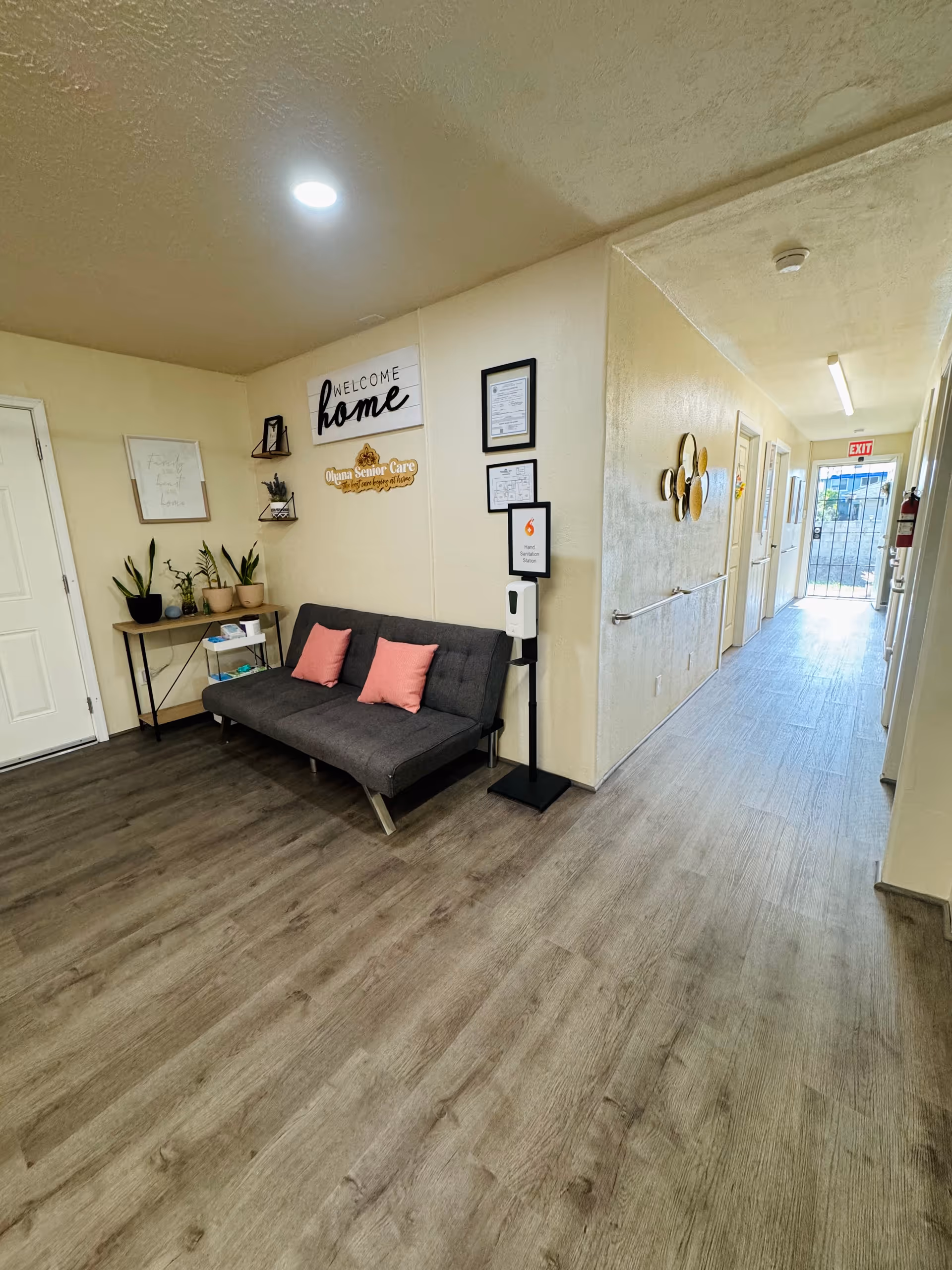 A hallway in a senior care facility with wood-look flooring and cream-colored walls. On the left side, there is a dark gray couch with two pink pillows, a small table with potted plants, and wall decorations including a sign that says 'WELCOME home' and another sign for Ohana Senior Care. A hand sanitizer dispenser is mounted on a stand next to the couch. The hallway extends to a door with an exit sign above it and a fire extinguisher mounted on the wall.
