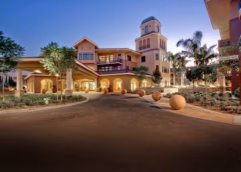 Entrance and front facade of a multi-story retirement community at dusk with a circular driveway, spherical bollards, palm trees and a tower.