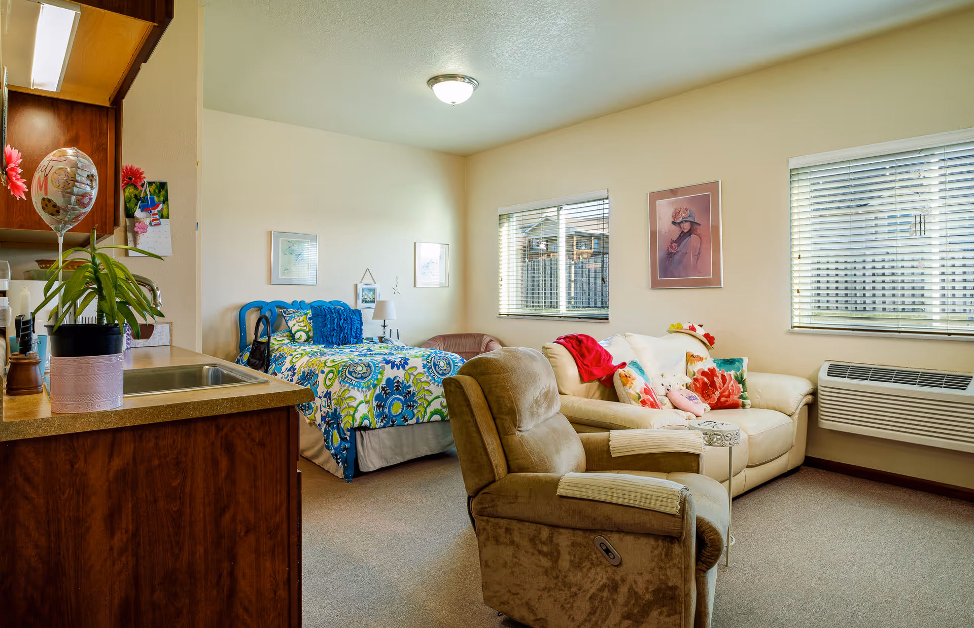A cozy room in a senior living facility featuring a single bed with colorful bedding, a beige reclining chair, and a cream-colored loveseat with floral pillows and a red throw. The room has two windows with blinds, a wall-mounted air conditioning unit, and a small kitchenette area with a sink and wooden cabinets. There are framed pictures on the walls and a potted plant on the counter.