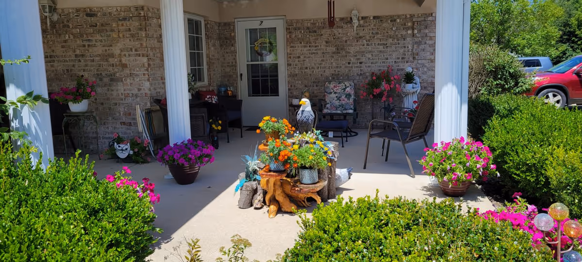 A covered patio area with brick walls and white columns, decorated with various potted flowers and plants. There are several chairs with cushions arranged around the patio, and a small table with a decorative eagle statue and more flowers in the center. Green bushes and colorful flowers surround the patio, and a red vehicle is partially visible in the background.