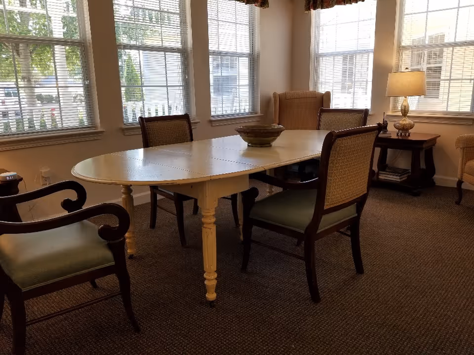 Oval dining table with several upholstered chairs in a sunlit common room with large windows and a lamp.