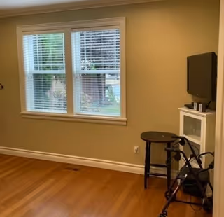 A small room with light brown walls and wooden floor. There is a window with white blinds letting in natural light. In the corner, there is a small white cabinet with a flat-screen TV on top, a round wooden stool, and a black walker nearby.