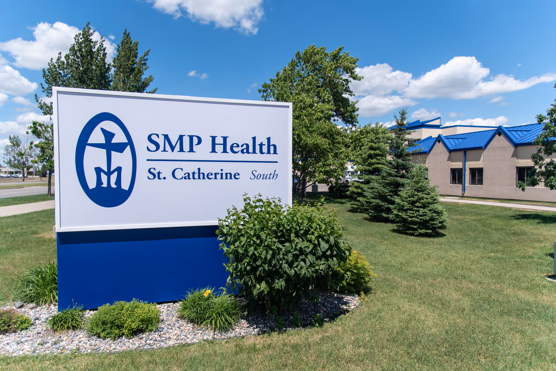 A large SMP Health St. Catherine South sign stands on a grassy lawn in front of a low building under a blue sky.