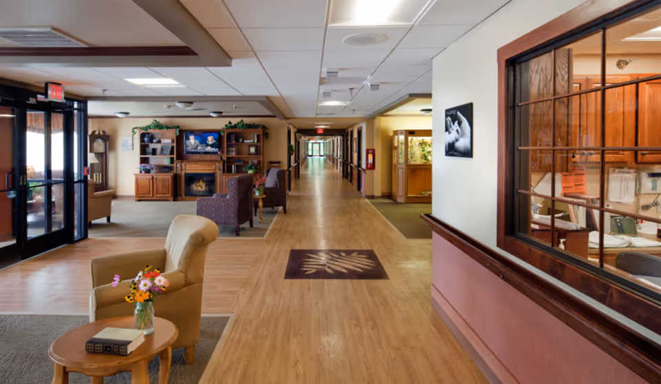 Interior view of a senior living facility hallway with wooden flooring, beige walls, and ceiling lights. There are comfortable armchairs and a small table with a vase of flowers and a book in the foreground. In the background, there is a seating area with armchairs, a wooden entertainment center with a TV showing a news program, and a fireplace. A large window with wooden framing looks into an office area on the right side. The hallway extends into the distance with handrails along the walls.