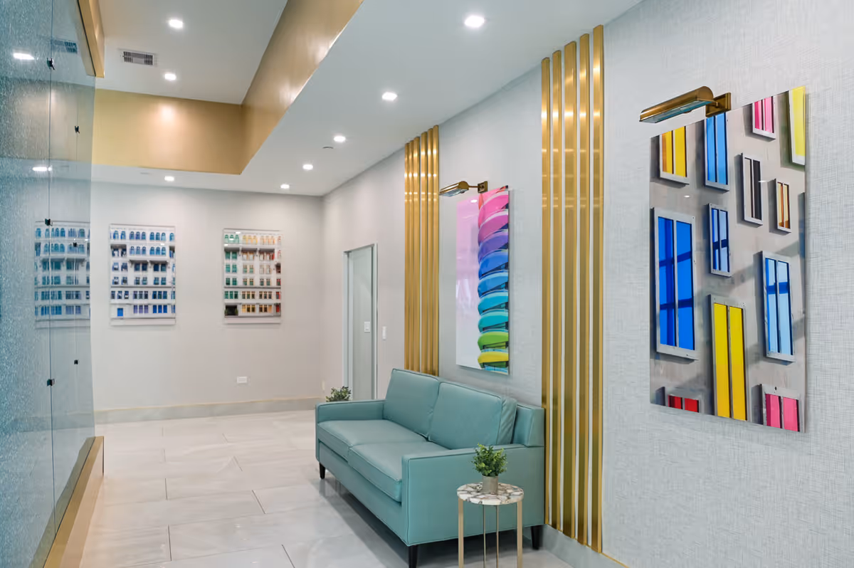 A modern interior waiting area with a light blue sofa and a small round side table with a plant. The walls are decorated with colorful abstract artwork and vertical gold accents. The floor is tiled in light colors, and the ceiling has recessed lighting.