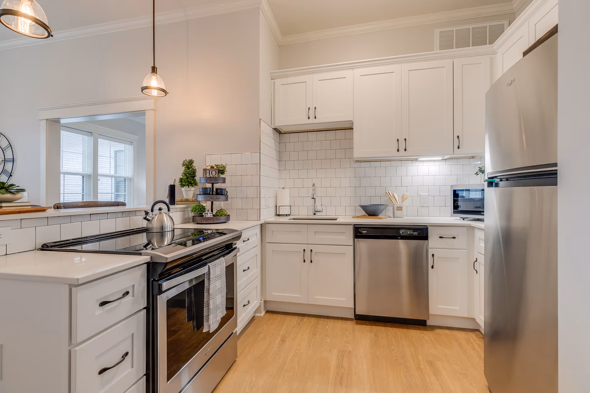Modern kitchen with white cabinets, stainless steel appliances including a refrigerator, dishwasher, and stove with a kettle on top. The backsplash is white tile, and there are wooden utensils and small potted plants on the countertops. Pendant lights hang from the ceiling, and a window with blinds is visible in the adjacent room.