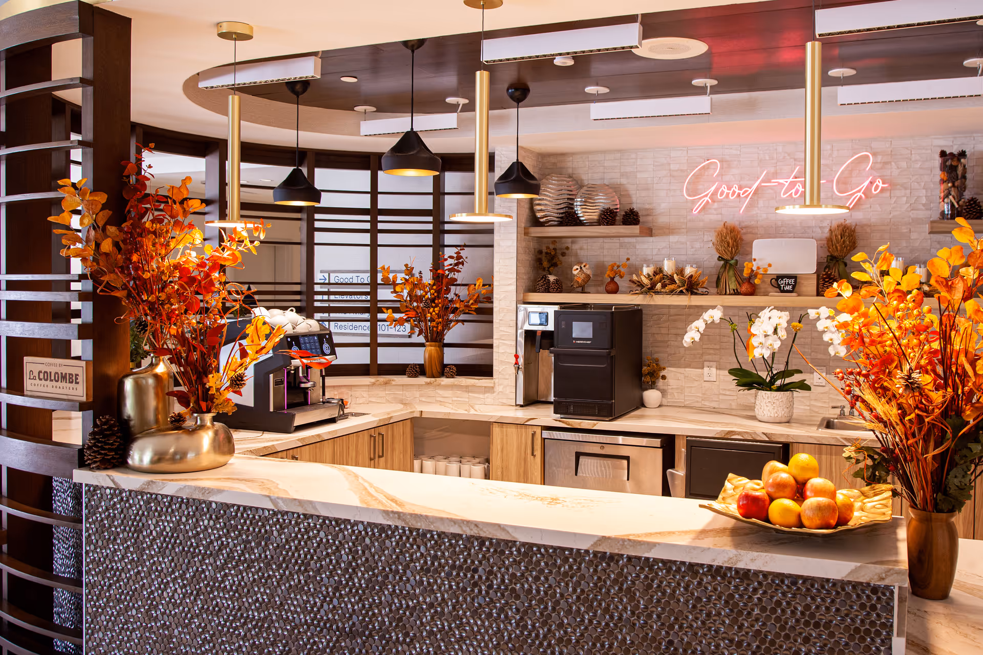 A modern coffee and snack bar area with a marble countertop, decorated with autumn-themed floral arrangements and a bowl of apples. Behind the counter are coffee machines, a sink, and shelves with decorative items including pine cones and vases. A neon sign on the wall reads 'Good to Go'. The space is warmly lit with pendant lights hanging from the ceiling.