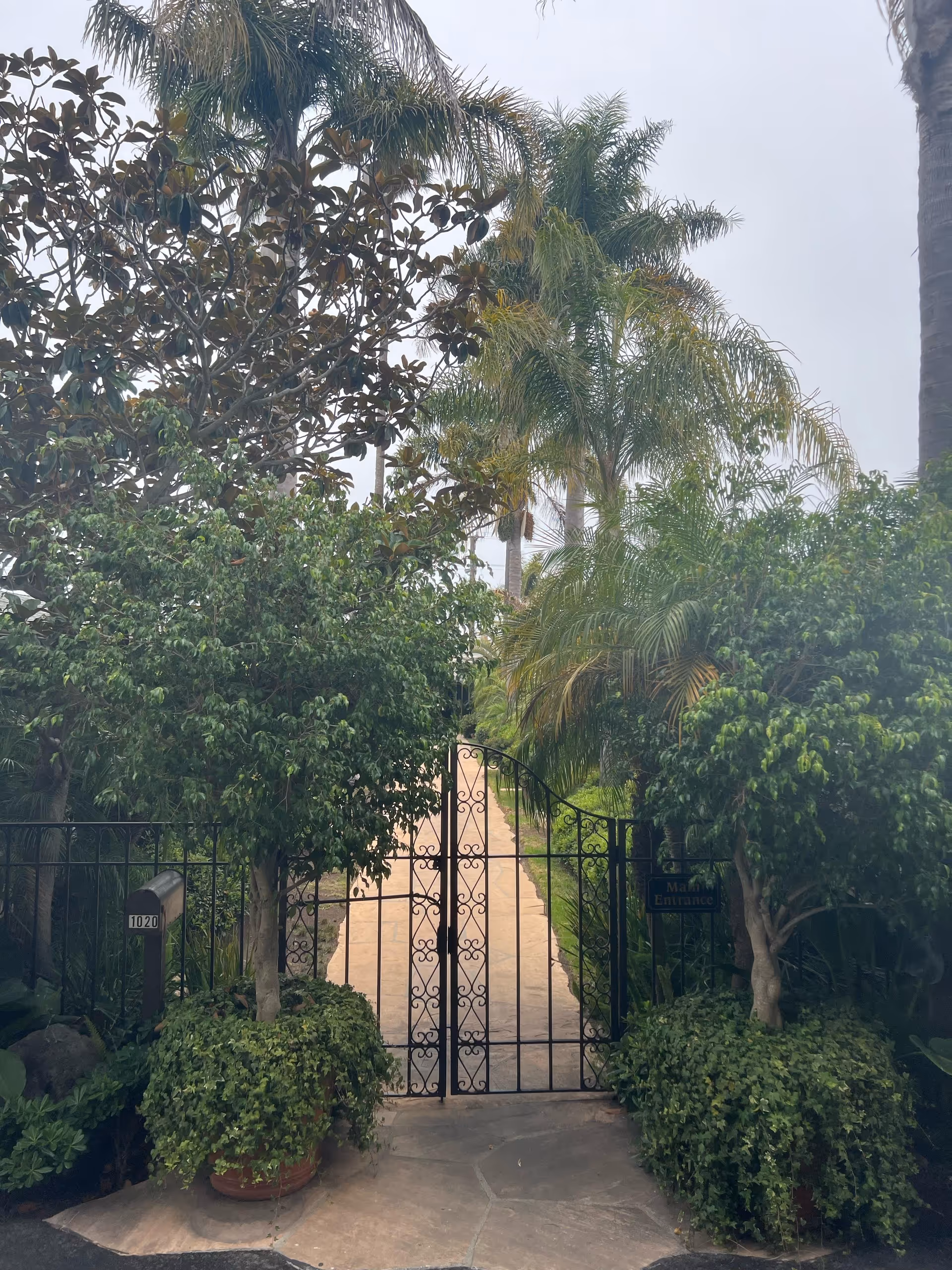 A black wrought iron gate with decorative scrollwork at the entrance of a pathway lined with lush green plants and palm trees on both sides. There are two potted plants with green foliage in front of the gate, and a mailbox labeled 1020 is visible on the left side. A small sign on the right side of the gate reads 'Main Entrance'.