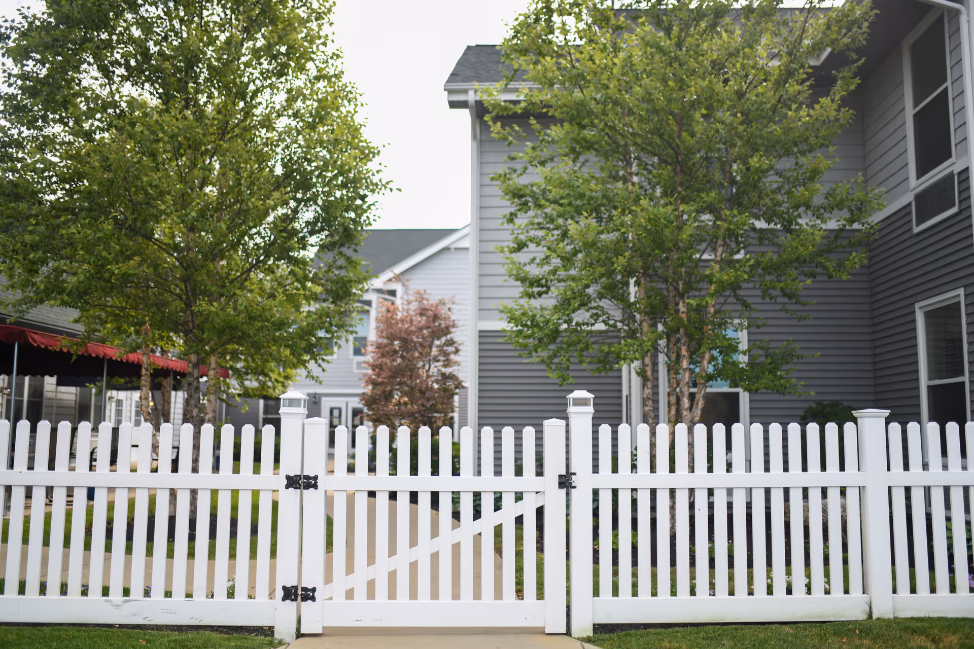 View of a white picket fence gate with a concrete pathway leading through it, surrounded by green grass and trees, with gray residential buildings in the background under an overcast sky.