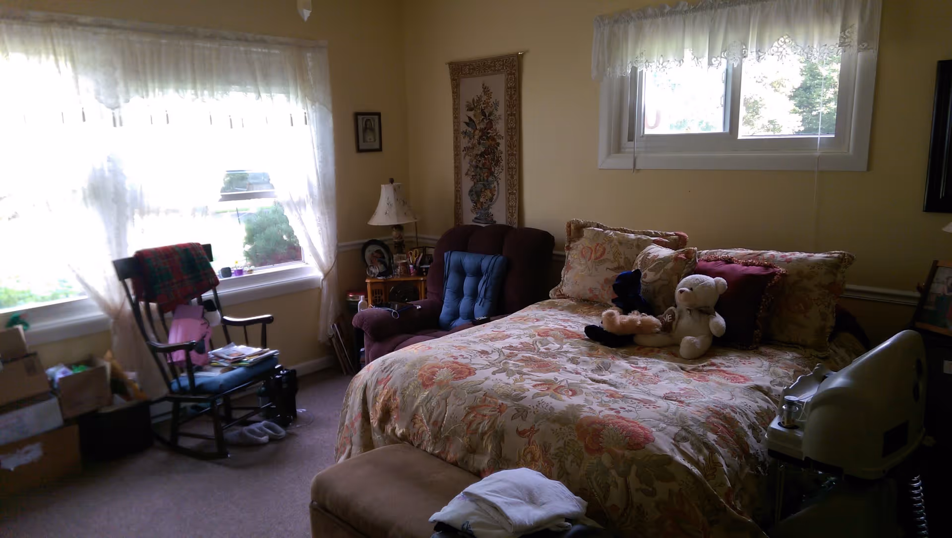 A cozy bedroom with a large bed covered in a floral patterned bedspread and several pillows, including stuffed animals. There is a cushioned armchair next to a small wooden side table with a lamp and framed photos. A rocking chair with a plaid blanket is positioned near two large windows with sheer curtains letting in natural light. Various personal items and boxes are visible around the room.