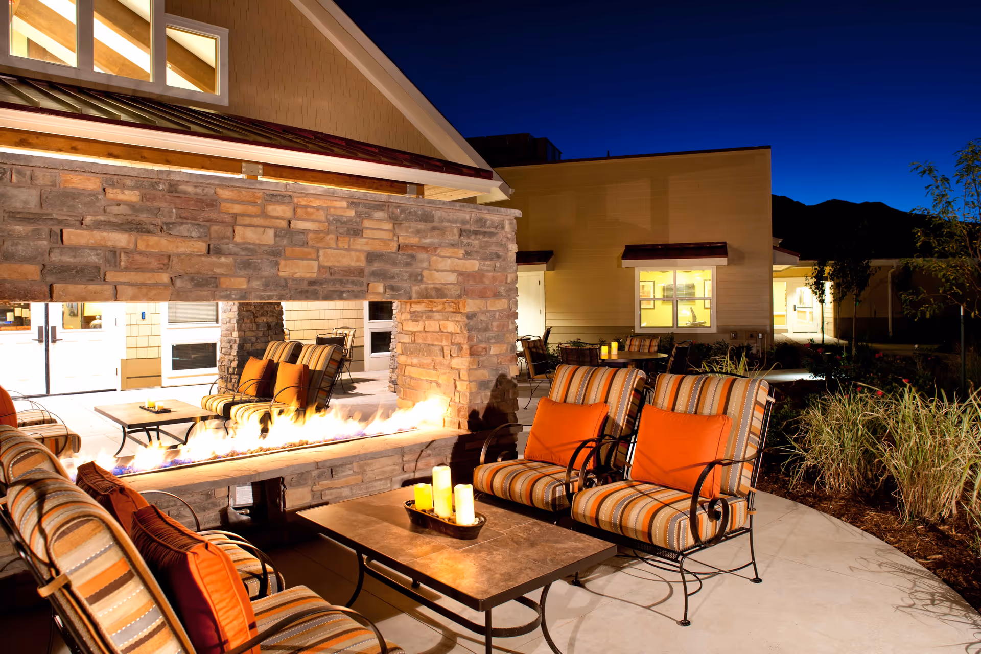 Outdoor seating area at night with cushioned chairs featuring orange and striped pillows around a rectangular fire pit table. The area is surrounded by a stone wall with a built-in fire feature and is part of a well-lit building exterior with windows and plants nearby.