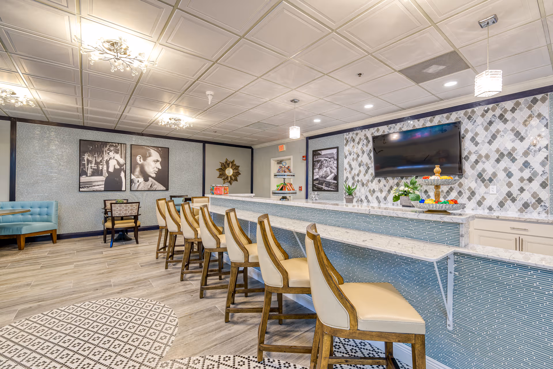 Communal dining and bar area in a senior living facility with a long counter and stools, wall-mounted TV, and lounge seating.