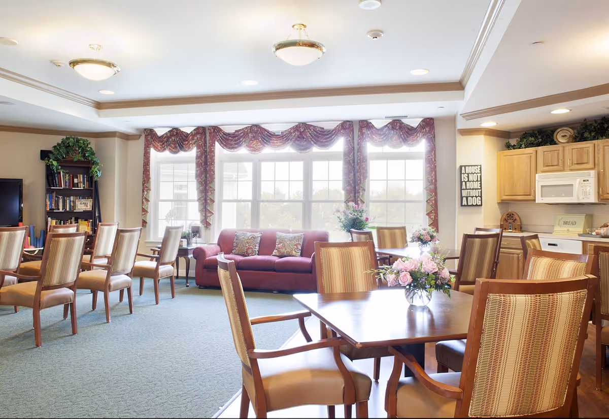A bright and spacious common area in a senior living facility featuring multiple wooden tables and chairs with striped upholstery. A red sofa with decorative pillows is positioned in front of large windows with patterned valances. The room includes a bookshelf with plants and books, a television, and a kitchenette with wooden cabinets, a microwave, and a dishwasher. Floral arrangements decorate the tables, and a wall sign reads 'A house is not a home without a dog.'